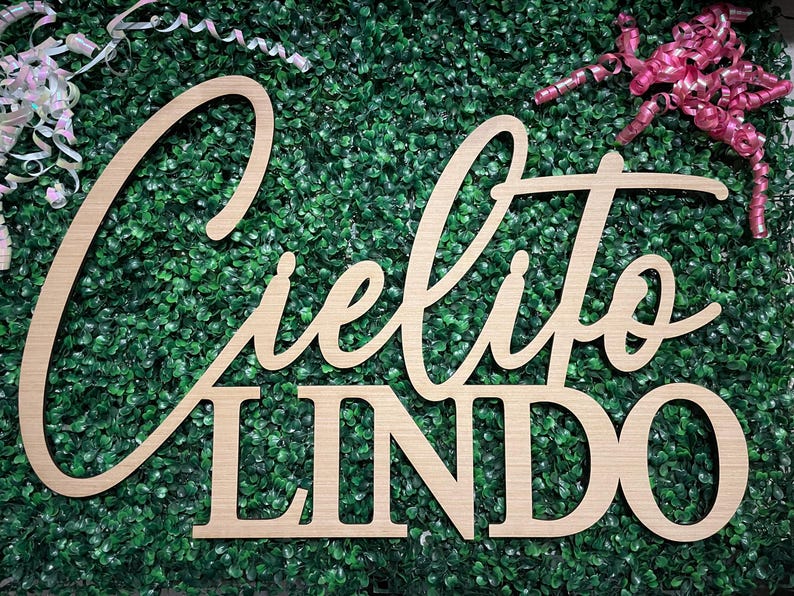 May include: Wooden sign with the words "Cielito Lindo" in a cursive font and block letters. The sign is made of light-colored wood and is set against a backdrop of green foliage. Decorative ribbons are in the background.