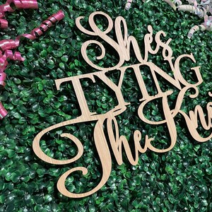 Puede incluir: Letrero de madera con la frase "She's Tying the Knot" en fuente cursiva. El letrero es de madera clara y est&aacute; colocado sobre un fondo de follaje verde y cintas rizadas rosas y blancas.