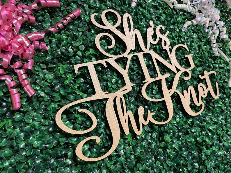 Puede incluir: Letrero de madera con la frase "She's Tying the Knot" en fuente cursiva. El letrero es de madera clara y est&aacute; colocado sobre un fondo de follaje verde y cintas rizadas rosas y blancas.