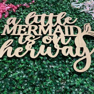May include: Wooden sign with the phrase "a little mermaid is on her way" in a cursive font. The sign has a mermaid tail design and is set against a green backdrop with pink and iridescent ribbons.
