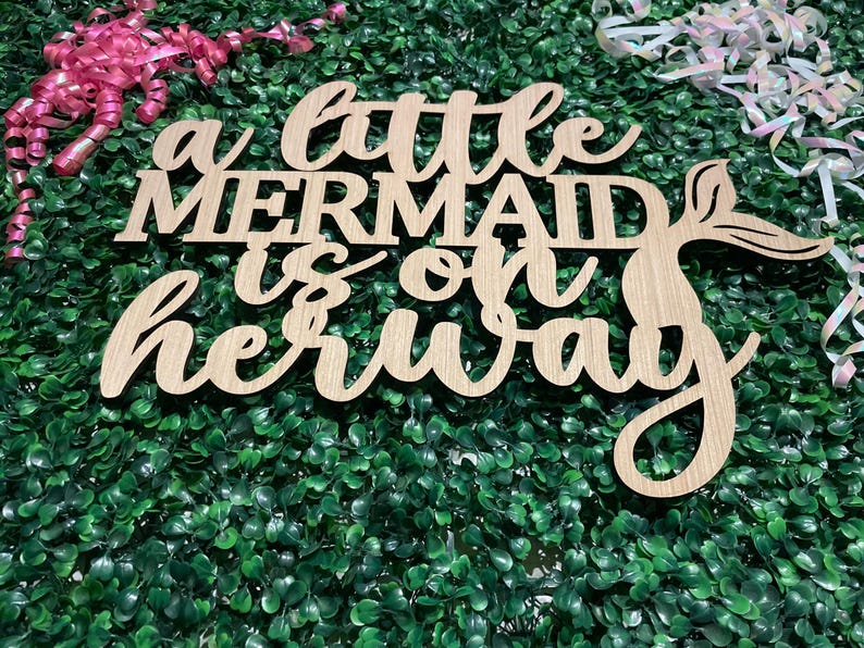 May include: Wooden sign with the phrase "a little mermaid is on her way" in a cursive font. The sign has a mermaid tail design and is set against a green backdrop with pink and iridescent ribbons.