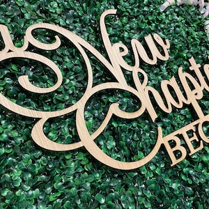 May include: A wooden sign with the words "A New Chapter Begins" in a cursive font. The sign is light brown and set against a backdrop of green foliage. Decorative ribbons are visible in the background.