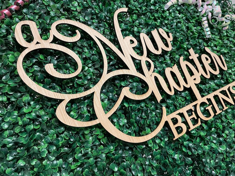 May include: A wooden sign with the words "A New Chapter Begins" in a cursive font. The sign is light brown and set against a backdrop of green foliage. Decorative ribbons are visible in the background.
