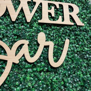 May include: Wooden sign with the words "Flower Bar" in a cursive font, set against a backdrop of green artificial foliage.