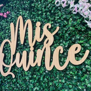 Op de afbeelding: Een lichtbruin houten bord met de woorden Mis Quince geschreven in een cursief lettertype. Het bord is geplaatst tegen een achtergrond van groen kunstblad met roze en iriserend witte krullende linten bevestigd aan de bovenhoeken.