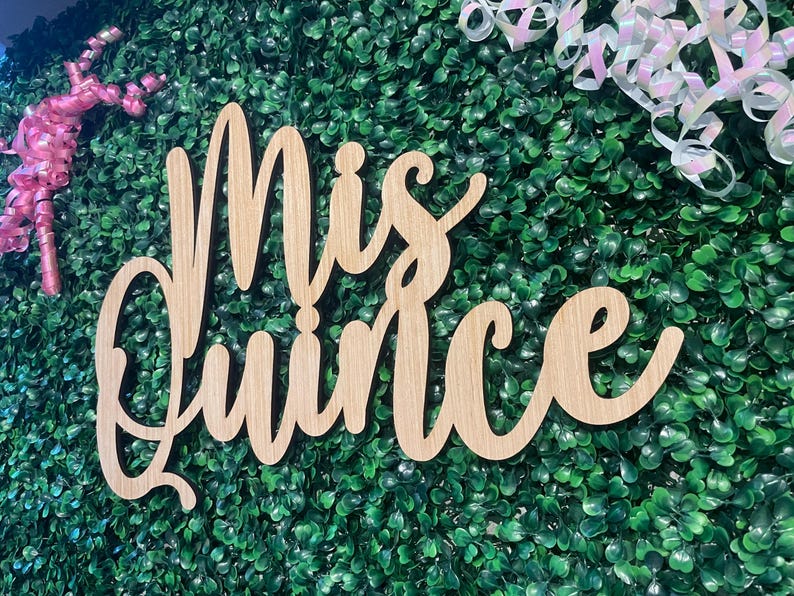 Op de afbeelding: Een lichtbruin houten bord met de woorden Mis Quince geschreven in een cursief lettertype. Het bord is geplaatst tegen een achtergrond van groen kunstblad met roze en iriserend witte krullende linten bevestigd aan de bovenhoeken.