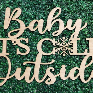 May include: Wooden sign with the text "Baby It's Cold Outside" in cursive font. The sign is decorated with a snowflake.