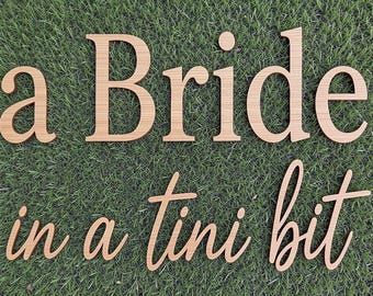May include: Light brown wooden cutout letters spelling the phrase a Bride in a tini bit. The top line features a serif font for a Bride, while the bottom line displays a cursive script font for the words in a tini bit, all with a wood grain.