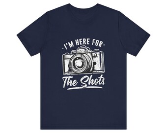 May include: Navy blue t-shirt with a white graphic of a vintage camera and the text "I'm Here For The Shots." The design features a detailed illustration of a camera, perfect for photography enthusiasts.