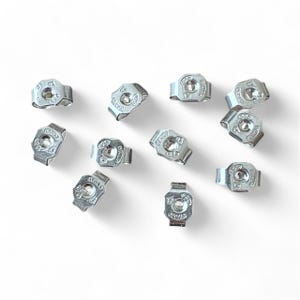 May include: A collection of silver-colored earring backs, scattered on a white surface. Each back has a small, circular design in the center and the word "ITALY" is visible on each back. The earring backs are made of metal.