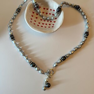 Peut inclure: Un collier de perles argentées avec des accents noirs et un pendentif en argent. Le collier est sur un plat blanc avec des cœurs rouges.