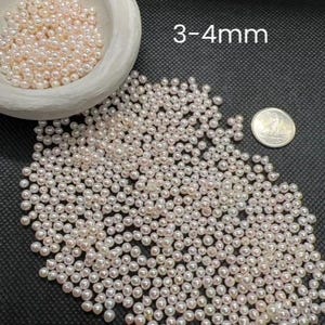 May include: A close-up image of a pile of small, white, and pink pearls. The pearls are approximately 3-4 mm in size. The pearls are scattered on a black surface.