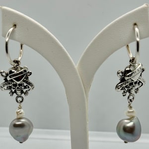 May include: Silver hoop earrings with a skull and crossbones charm and a grey pearl drop. The charm features the words "Love Kills Slowly".