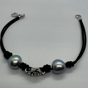 May include: A black leather bracelet with two iridescent grey pearls and a silver rose charm. The bracelet has a clasp closure and is approximately 18 cm in length.
