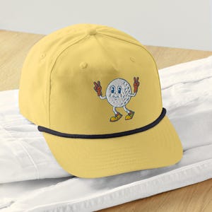 May include: A yellow baseball cap with a black rope trim and an embroidered cartoon golf ball with a smiling face and peace signs.