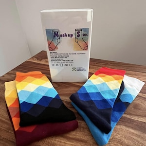 May include: A selection of colourful socks with a geometric pattern in shades of red, orange, yellow, blue, and black are displayed next to a white box. The box has the text "Mash up Sox" and washing instructions. The socks are made of a cotton, polyester, and spandex blend.