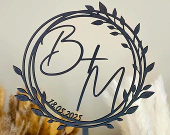 May include: Black metal cake topper with a wreath design and the initials "B + M" and the date "18.05.2025".