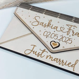 May include: A wooden sign with a white background and black border. The sign is shaped like an envelope and has the text "Saskia & Fra 02.05.2025" and "Just married" written on it. There is a small wooden heart in the upper right corner of the sign.