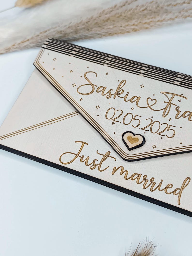 May include: A wooden sign with a white background and black border. The sign is shaped like an envelope and has the text "Saskia & Fra 02.05.2025" and "Just married" written on it. There is a small wooden heart in the upper right corner of the sign.