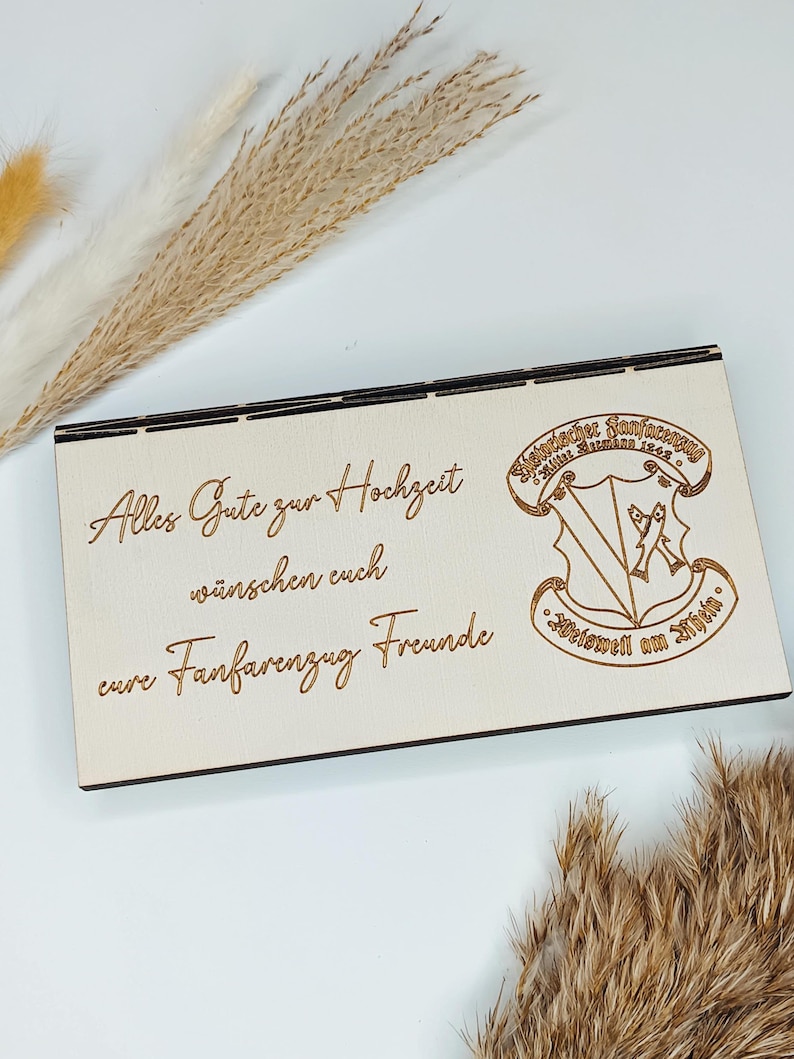May include: A wooden sign with the text "Alles Gute zur Hochzeit" and "eure Fanfarenzug Freunde" in a cursive font. The sign also features a crest with the text "M&uuml;nchner Fanfarenzug" and "Mittenwald 1924" in a traditional font.