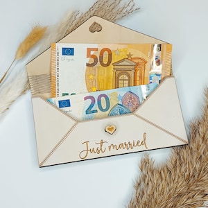 May include: A wooden envelope with a heart cutout and the words "Just married" engraved on the front. The envelope is open and contains two Euro bills, a 20 Euro bill and a 50 Euro bill.