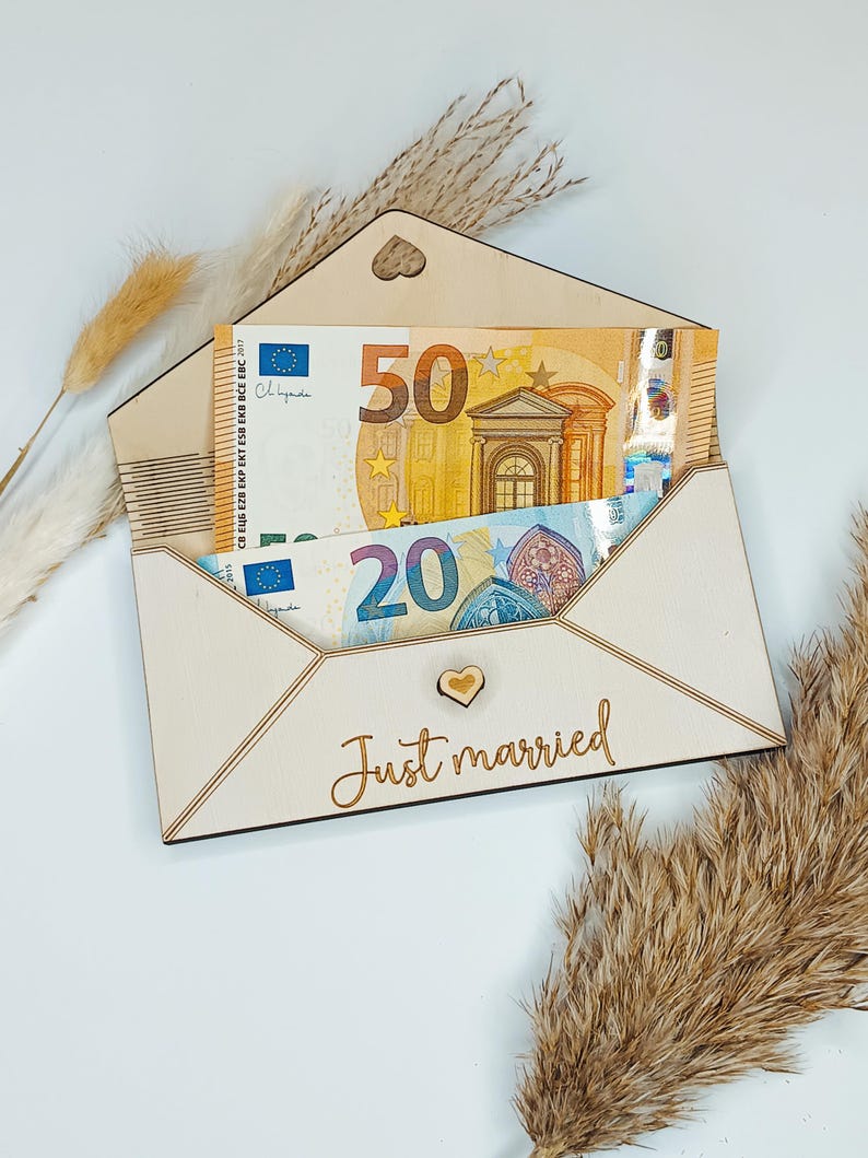 May include: A wooden envelope with a heart cutout and the words "Just married" engraved on the front. The envelope is open and contains two Euro bills, a 20 Euro bill and a 50 Euro bill.
