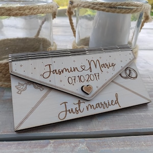 May include: A wooden envelope with the text "Jasmin & Mario 07.10.2017 Just married" engraved on it. The envelope is decorated with a heart, two birds, and two rings.