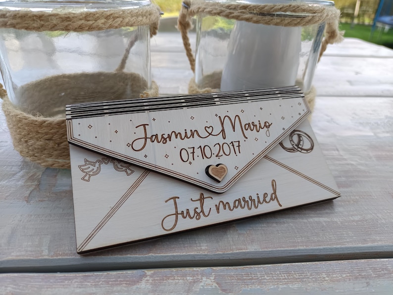 May include: A wooden envelope with the text "Jasmin & Mario 07.10.2017 Just married" engraved on it. The envelope is decorated with a heart, two birds, and two rings.