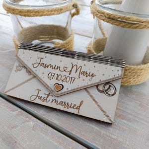 May include: A wooden envelope with the text "Jasmin & Mario 07.10.2017 Just married" engraved on it. The envelope is decorated with a heart and two rings.