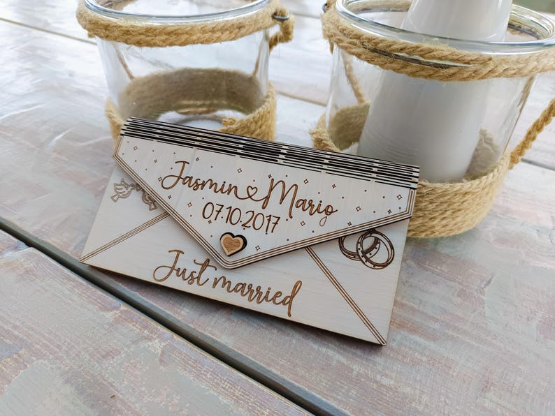 May include: A wooden envelope with the text "Jasmin & Mario 07.10.2017 Just married" engraved on it. The envelope is decorated with a heart and two rings.