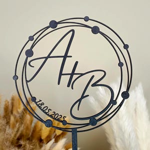 Könnte beinhalten: Schwarzer Holz-Kuchentopper mit den Initialen "A & B" in einer kursiven Schriftart innerhalb eines Kreises mit einem gepunkteten Linienmuster. Das Datum "18.05.2025" steht unter den Initialen.