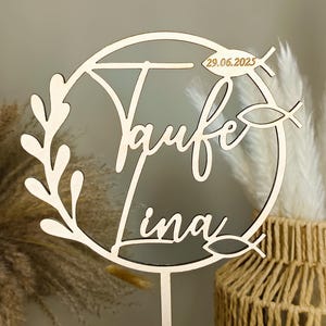 Könnte beinhalten: Ein Kuchenaufsatz aus Holz mit den Worten "Taufe Lina" und dem Datum "29.06.2025" ausgeschnitten. Das Design umfasst einen kreisförmigen Rahmen, Fischsymbole und einen Blätterzweig. Der Aufsatz ist aus hellem Holz gefertigt.