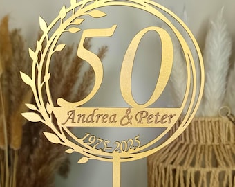 May include: A gold-colored wooden cake topper with the number 50 in a circular design, surrounded by leaves. The names "Andrea & Peter" and the years "1975-2025" are also displayed. The topper is on a stick.