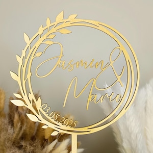 Könnte beinhalten: Goldener Holz-Kuchentopper mit einem floralen Kranzdesign. Der Topper zeigt die Namen "Jasmin & Mario" und das Datum "03.05.2025".