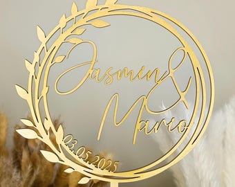 May include: Gold wooden cake topper with a floral wreath design. The topper features the names "Jasmin & Mario" and the date "03.05.2025".