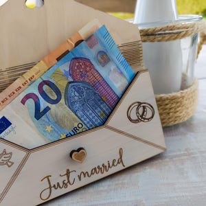 May include: A wooden envelope with a slot for money, decorated with the words "Just married" and two rings. The envelope is filled with a twenty euro bill.