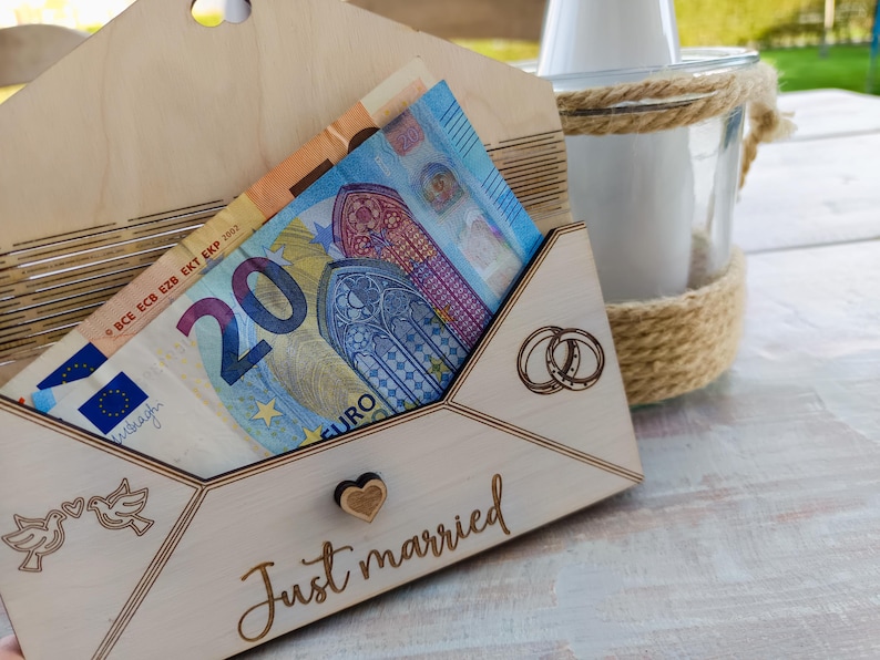 May include: A wooden envelope with a slot for money, decorated with the words "Just married" and two rings. The envelope is filled with a twenty euro bill.