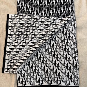 May include: A folded white scarf with a black repeating pattern. The scarf has a black border and is made of a soft material. The pattern is a repeating logo design.