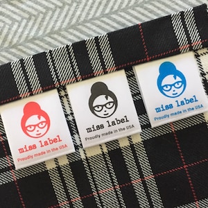 May include: Three white fabric labels with a black and white plaid background. Each label features a cartoon woman with glasses and a bun. The text on the labels reads "miss label" and "Proudly made in the USA".