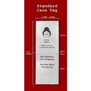 May include: A white rectangular fabric care tag measuring 1 5/16 inches wide by 3 1/2 inches long. The front features a logo with a person wearing glasses, the text Miss Label, and Made in the USA of imported materials. The back lists 80% Bamboo, 15% Polyester, Machine Wash, and Tumble Dry.