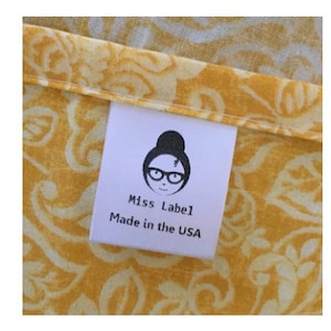 May include: A white fabric clothing label sewn onto yellow fabric with a white floral pattern. The label features a black graphic of a person with a bun hairstyle and glasses, with the text Miss Label and Made in the USA printed below it.