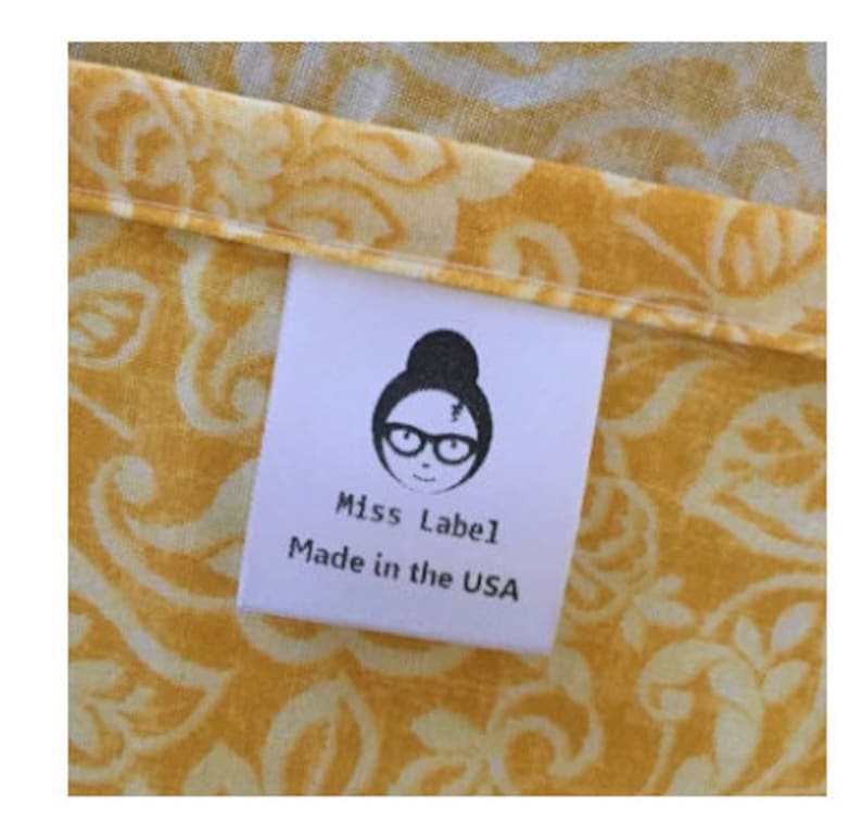 May include: A white fabric clothing label sewn onto yellow fabric with a white floral pattern. The label features a black graphic of a person with a bun hairstyle and glasses, with the text Miss Label and Made in the USA printed below it.