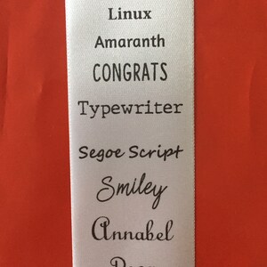 May include: A white rectangular fabric label featuring a list of black printed text in various fonts: Franklin Gothic, Linux, Amaranth, CONGRATS, Typewriter, Segoe Script, Smiley, Annabel, Rosa, and the word NOTES enclosed in individual square boxes.