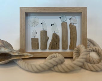 May include: A framed artwork featuring glass birds perched on driftwood. The birds are white with black beaks, and the driftwood is various shades of brown. A sand dollar is also included in the frame. A seashell and rope are in the foreground.
