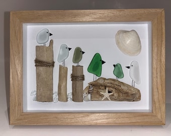 May include: A framed artwork featuring glass bird figures in white, green, and gray perched on wooden posts and driftwood. A seashell and starfish are also included in the beach-themed art piece. The frame is made of light-colored wood.