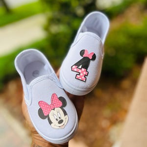 Puede incluir: Zapatillas blancas de lona para ni&ntilde;os peque&ntilde;os. Un zapato presenta un gr&aacute;fico de Minnie Mouse con un lazo rosa de lunares, y el otro muestra el n&uacute;mero "4" con un acento rosa de lunares. Los zapatos est&aacute;n dise&ntilde;ados para ni&ntilde;os.