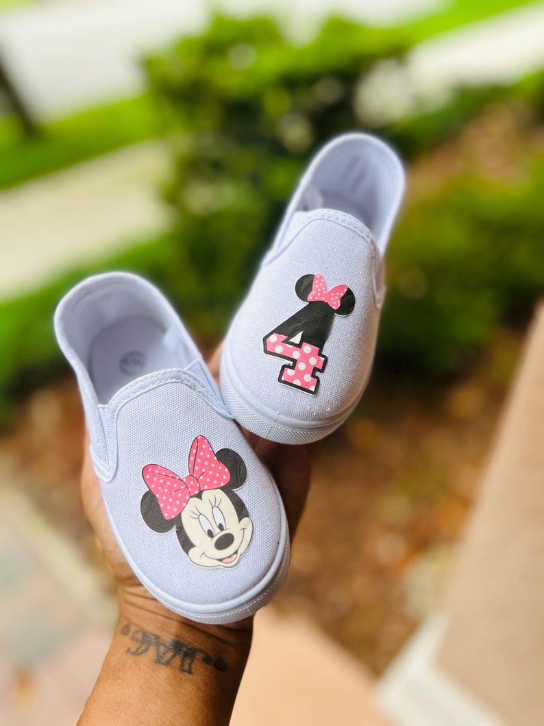 Puede incluir: Zapatillas blancas de lona para ni&ntilde;os peque&ntilde;os. Un zapato presenta un gr&aacute;fico de Minnie Mouse con un lazo rosa de lunares, y el otro muestra el n&uacute;mero "4" con un acento rosa de lunares. Los zapatos est&aacute;n dise&ntilde;ados para ni&ntilde;os.