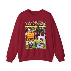 May include: A maroon sweatshirt featuring a football action shot with the text "AIR JAYDEN." The image shows a player in a black uniform jumping over other players on a green field. The sweatshirt is a solid maroon color.