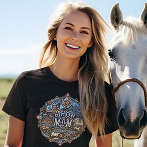 May include: A woman with long blonde hair smiles while standing next to a white horse. She is wearing a black t-shirt with a colorful graphic design that says "Show Mom".
