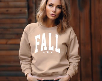Puede incluir: Sudadera beige con la palabra "FALL" en letras blancas grandes y tres estrellas blancas debajo. La sudadera es de color neutro y tiene mangas largas. La persona lleva vaqueros azules.